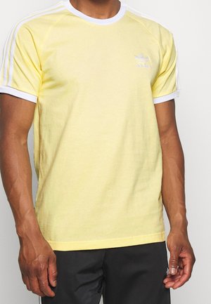 Mężczyzna w jasnopomarańczowej koszulce Adidas z białymi paskami na ramionach i rękawach, w połączeniu z czarnymi spodniami Adidas.