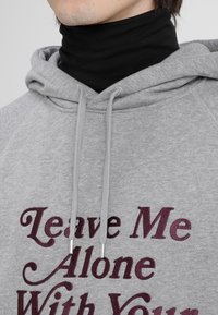 Grå hoodie med en svart polotröja under. Har texten "Leave Me Alone With Your" i bordeauxröd färg. Dragsnören med metallspetsar.