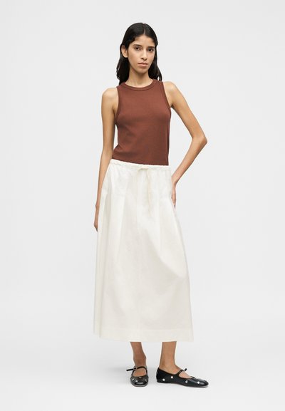 Καφέ αμάνικη μπλούζα σε συνδυασμό με μια μακριά, λευκή, πλισέ φούστα. Το μοντέλο φοράει μαύρα, επίπεδα παπούτσια με διακοσμητικά στοιχεία. Απλός και μινιμαλιστικός σχεδιασμός.