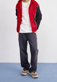 Červeno-černá větrovka s bílými akcenty, bílé tričko, tmavě šedé džíny a vícebarevné tenisky. Ležérní sportovní vzhled.