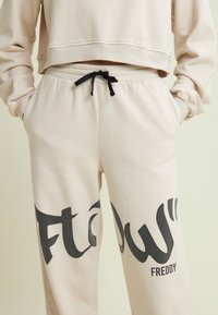 Pantalones de chándal beige con cordón negro, que presentan un gran texto en gris "FLOW" y el nombre de la marca "FREDDY". Tela suave, corte relajado, cintura elástica.