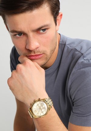 Jonge man in grijs overhemd die zijn kin op zijn hand laat rusten, draagt een gouden metalen horloge met een ronde wijzerplaat en Romeinse cijfers.