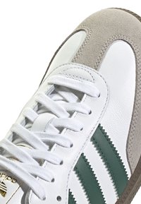 Vita lädersneakers med gröna ränder, en grå mockadetalj och textilskosnören. Guldlogotyp nära skosnörena. Strukturerad gummisula.