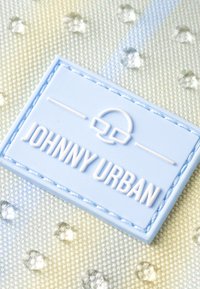 Világoskék, téglalap alakú címke, dombornyomott "JOHNNY URBAN" felirattal, steppelt szélekkel, csillogó, texturált háttérrel és apró, kerek díszítőelemekkel.