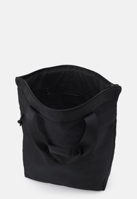 Sac fourre-tout noir en tissu texturé, avec deux poignées, un haut zippé et une poche intérieure. Design minimaliste avec un logo à l'avant.