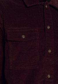 Burgundy kordfløyelskjorte med knappedown-krage, som har to brystlommer og brune knappdetaljer längs placket.