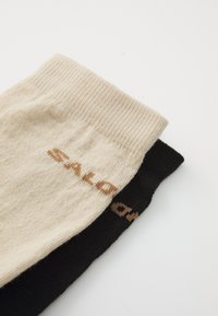 Two socks, one beige and one black, with a smooth texture. Each features the word "Salomon" in contrasting colors near the cuff.