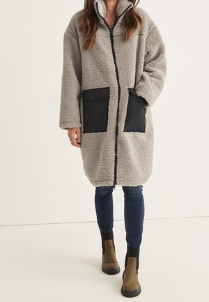 Femme portant un long manteau sherpa beige avec des poches noires, un jean bleu et des bottines marron, debout devant un fond uni.