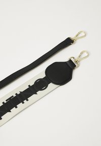 Pulseira de couro preta com ferragens em tom de ouro, apresentando lona branca com letras pretas em negrito e um destaque em couro texturizado.