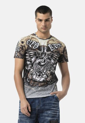 Jonge man met kort haar die een grafisch T-shirt draagt met een motorfiets motor, vleugels, schedels en tekst, gecombineerd met blauwe spijkerbroek.