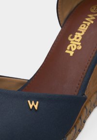 Prim-plan al unei sandale albastre cu platformă, talpă împletită și un emblamă aurie în formă de "W" pe breteaua din față.
