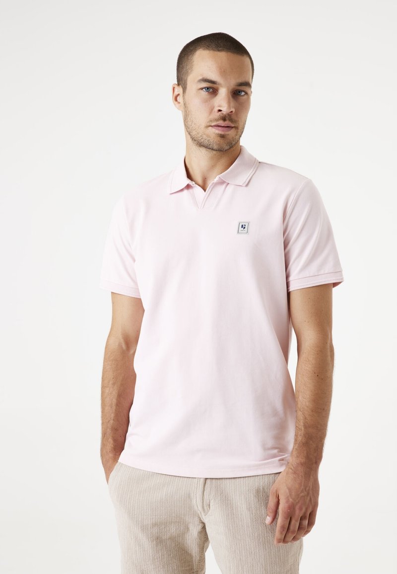 Camisa tipo polo rosa claro con cuello, mangas cortas y un pequeño logo cuadrado en el lado izquierdo del pecho. Material de algodón suave, diseño ajustado.