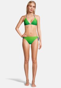 Vrouw die blootsvoets staat en een groene gestreepte string bikini met blauwe banden draagt, tegen een simpele witte achtergrond.