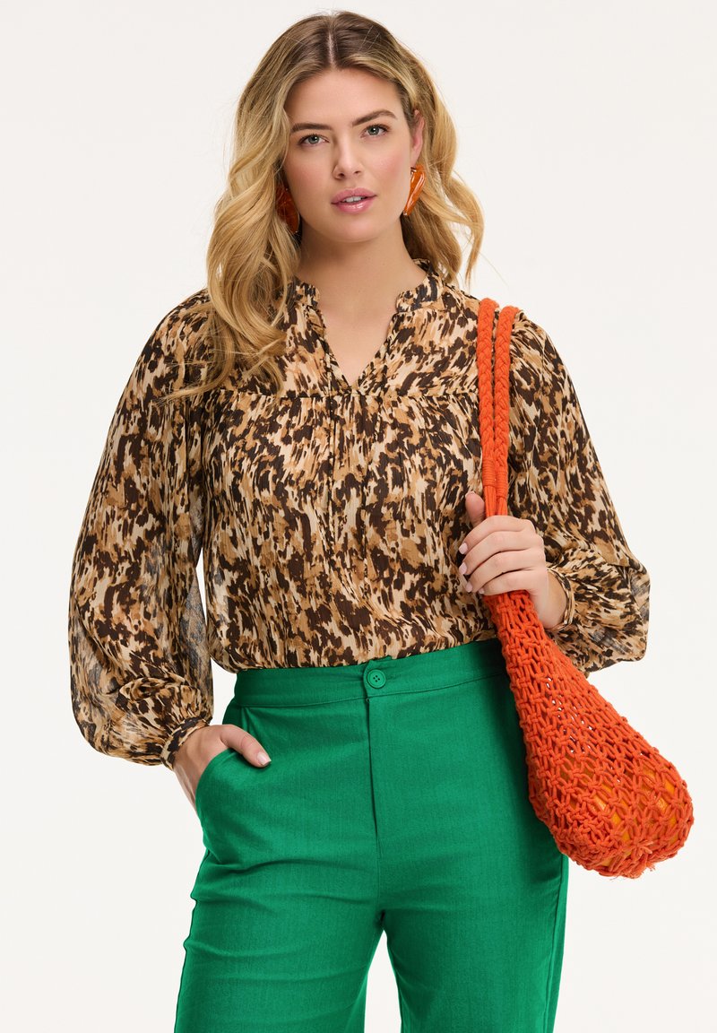 Blusa marrone e beige con motivo, maniche a sbuffo, abbinata a pantaloni verdi. In mano c'è una borsa tessuta arancione con un design texturizzato.