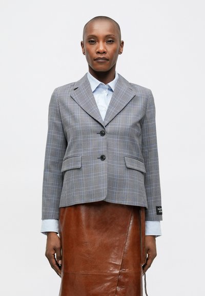 Blazer à carreaux gris avec deux boutons, poches avant et revers crantés. Porté sur une chemise bleu clair et associé à une jupe en cuir marron.