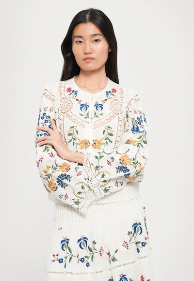 Weiße bestickte Bluse mit floralen Mustern in Blau, Gelb und Pink, mit einem Spitzenbesatz an der Passe und langen Ärmeln, kombiniert mit einem passenden Rock.