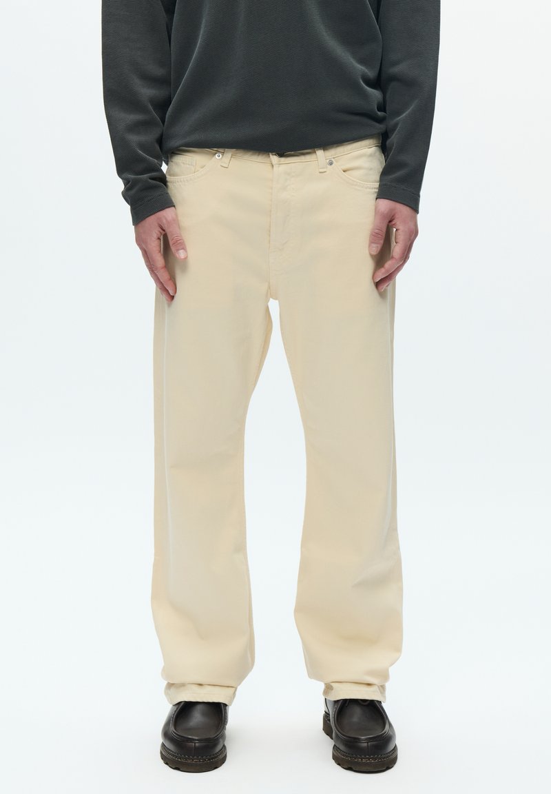 Béžové široké nohavice z bavlny s prednými vreckami, zipsom na zapínanie a kontrastným prešívaním, doplnené čiernymi topánkami.