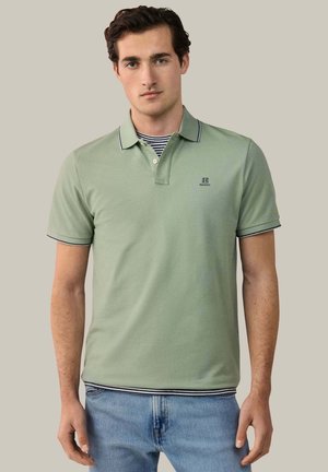 Jonge man die een lichtgroen poloshirt draagt over een gestreept shirt en een blauwe spijkerbroek, staand tegen een effen grijze achtergrond.