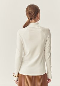 Cremfarbener Rollkragenpullover mit langen Ärmeln, ribbeliger Textur und einer taillierten Passform. Ergänzt durch einen gerüschten braunen Rock und Armbänder.