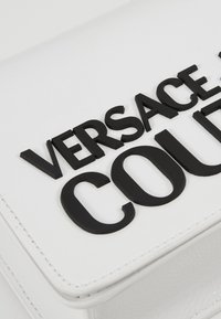 Valkoinen nahkainen käsilaukku, jossa on kohokuvioitu musta 'VERSACE COURT' -teksti edessä. Ominaisuuksina minimalistinen muotoilu ja siistit ompeleet.