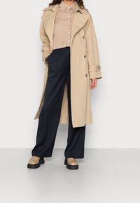 Beżowy trencz z podwójnym zapięciem, w połączeniu z paskowaną koszulą i granatowymi spodniami o szerokich nogawkach. Masywne beżowe buty na platformie.