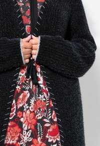 Mustat tekstuurilla varustetut neuletakit, joissa on valkoiset tikkaus, käytetään kukkakuvioisen punamustat mekon päällä. Mekossa on nyörikiinnitys ja tupsut.