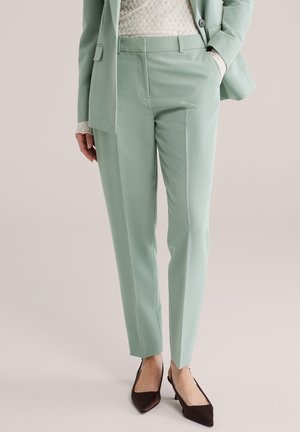 Frau trägt hellgrüne, maßgeschneiderte Hose und passenden Blazer, mit einem cremefarbenen Spitzen-Top und schwarzen spitzen Schuhen.