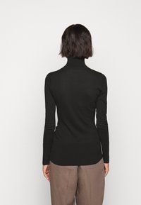 Frau mit kurzen dunklen Haaren, die einen schwarzen Rollkragenpullover und braune Hosen trägt, steht mit dem Rücken zu einem einfarbigen Hintergrund.