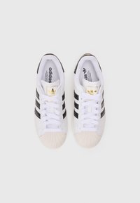 Sapatilhas brancas com riscas pretas, apresentando um upper texturizado, bico arredondado e detalhes em dourado no logo da língua. Atacadores planos e sola em borracha.