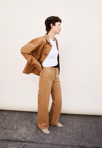 Veste beige avec des boutons noirs, t-shirt blanc et pantalon large marron clair. Tissu lisse, coupe décontractée et design minimaliste sans motifs.