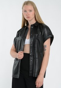 Sort læder skjorte med korte ærmer, frontlukning med knapper, to brystlommer og en glat, skinnende overflade. Bæres over en hvid tanktop.