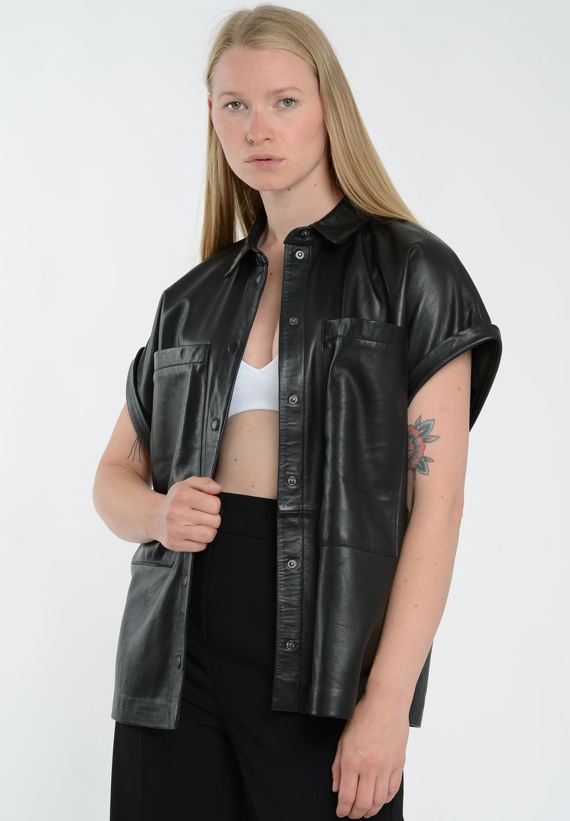 Sort læder skjorte med korte ærmer, frontlukning med knapper, to brystlommer og en glat, skinnende overflade. Bæres over en hvid tanktop.