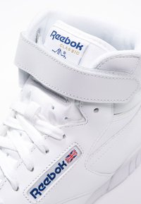 Baskets Reebok blanches avec un design montante, en cuir, bande Velcro, patch logo et texture de surface lisse.