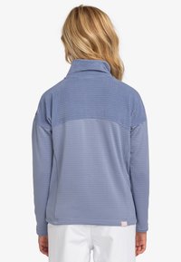 Polarowy pulower w jasnoniebieskim kolorze z teksturowanym, pikowanym wzorem. Posiada wysoki kołnierz i płaskie szwy. Po lewej stronie znajduje się detal z logo w pobliżu ramienia.