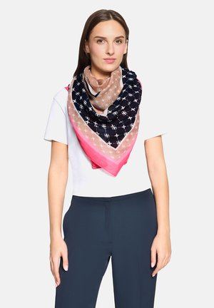 Donna che indossa una maglietta bianca a maniche corte, pantaloni blu navy e una grande sciarpa quadrata con motivi geometrici blu navy, beige e rosa annodata al collo.