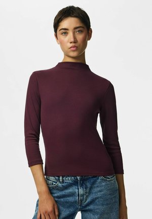 Jeune personne aux cheveux courts et foncés, portant un haut ajusté bordeaux à manches longues et un jean bleu sur un fond blanc uni.