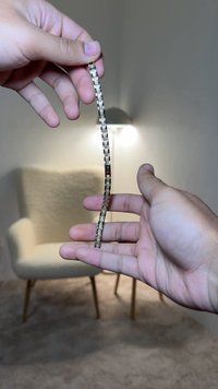 Brățară metalică cu linkuri alternative în nuanțe închise și deschise, design flexibil, ținută între degete pe un fundal neutru.