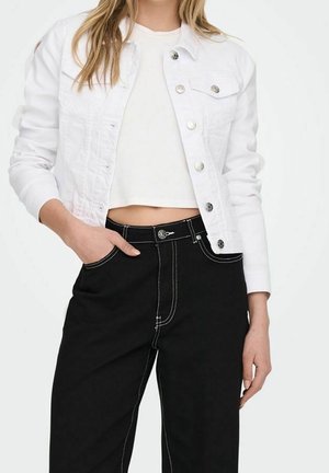 Vrouw draagt een wit cropped jasje over een wit crop top en hoge taille zwarte broek met witte stiksels, hand in de zak.
