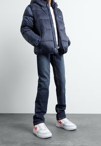 Doudoune bleu marine avec capuche, fermeture à glissière et rembourrage texturé, associée à un jean en denim bleu et des chaussures blanches avec un accent rouge.