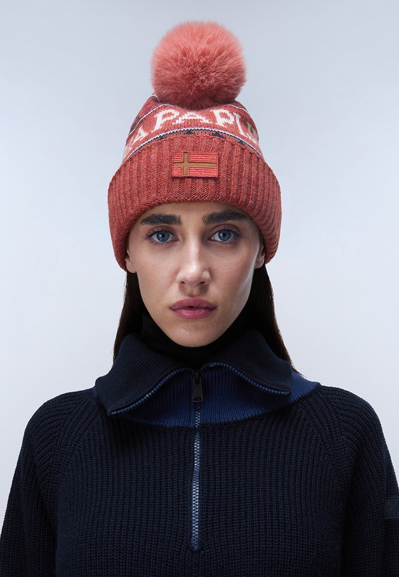 Berretto lavorato a maglia di colore rosso ruggine con un grande pom-pom. Presenta un motivo lungo il bordo e una toppa con una bandiera. Texture a coste.