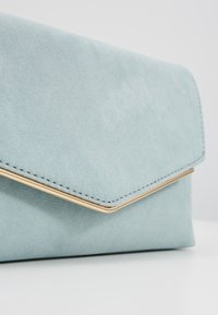 Světle modrá semišová kabelka s texturovaným povrchem, se zlatým akcentem na předním klopě a čistým, minimalistickým designem.