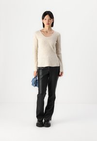Pull en tricot côtelé beige avec un décolleté en scoop, associé à un pantalon en faux cuir noir. Le mannequin tient un mini sac à main bleu.
