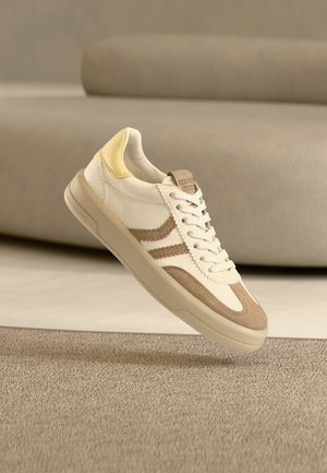 Witte en beige sneaker met een gele hielaccent, uitgevoerd in suède en glad leer, met platte veters en een rubberen zool.