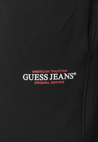 Juodos medvilninės sportinės kelnės su spausdintu logotipu ir raudona bei balta tekstu: "AMERIKOS TRADICIJOS GUESS JEANS ORIGINALŲ DIZAINAI."