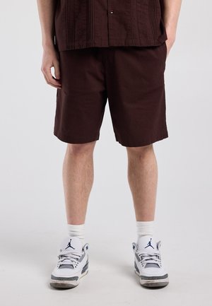 Persoon draagt donkerbruine korte broek, een bijpassend bruin shirt, witte sokken en witte en grijze sneakers met een blauw logo, staand op een lichte achtergrond.