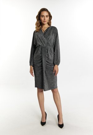 Metallisch silbernes Kleid mit langen Ärmeln, gerafftem Taillenbereich, knie-langer Saum und sanftem Schimmer. Kombiniert mit schwarzen High Heels.