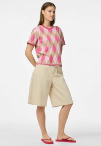 Růžový a béžový argyle pletený svetr s krátkým rukávem, v kombinaci s béžovými šortkami ke kolenům. Modelka má na sobě růžové flip-flopy.