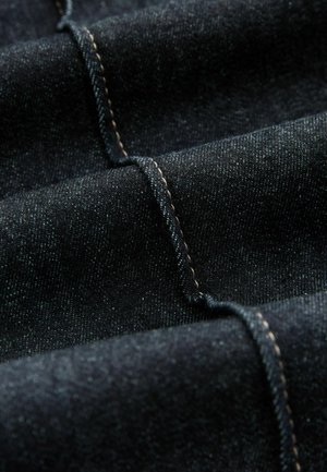 Ciemny dżins o subtelnej teksturze, z kontrastowym beżowym przeszyciem i gładkimi, złożonymi warstwami.