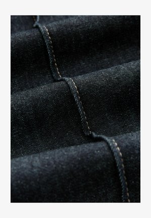 Ciemny dżins o subtelnej teksturze, z kontrastowym beżowym przeszyciem i gładkimi, złożonymi warstwami.