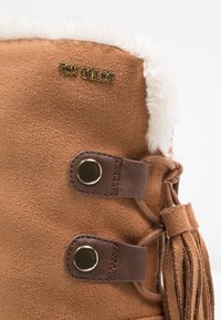 Hnědé suede kotníkové boty s bílým kožešinovým lemem, disponující zlatým logem, hnědými koženými akcenty a dvěma kovovými knoflíky. Dekorativní detail s třásněmi.
